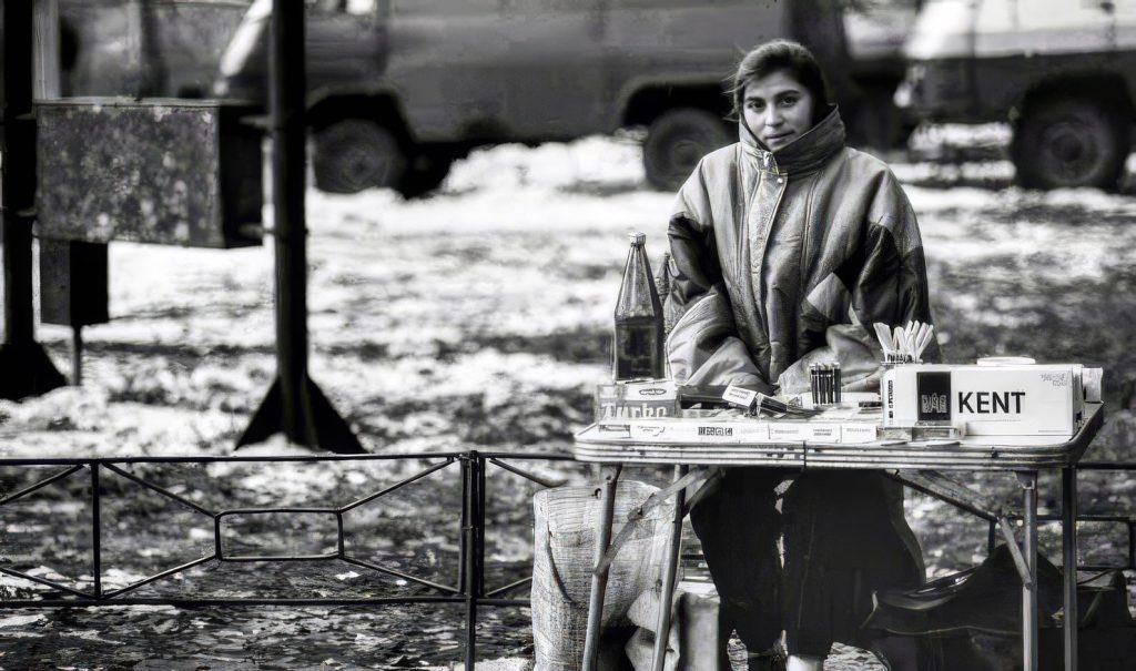 Strada urbana della Romania negli anni ’90 con bancarelle improvvisate e venditori ambulanti che offrono jeans, sigarette e profumi, simbolo del commercio di sopravvivenza dopo la caduta del comunismo.