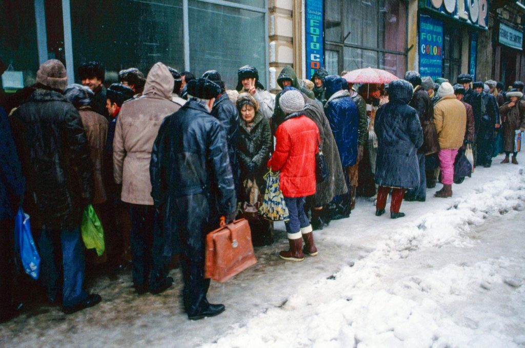Persone in coda davanti a un negozio durante la crisi economica in Romania dopo il 1989, simbolo della scarsità e delle difficoltà quotidiane.